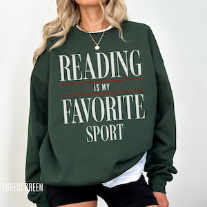 May include: A forest green sweatshirt featuring the text "READING IS MY FAVORITE SPORT" in white, with a red line separating the words. The sweatshirt is a crew neck style with long sleeves. The bottom of the sweatshirt is visible.