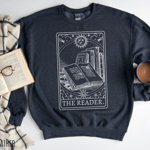 Puede incluir: Sudadera gris oscuro jaspeada con un gr&aacute;fico blanco de una carta de tarot que representa una pila de libros con el texto "THE READER."