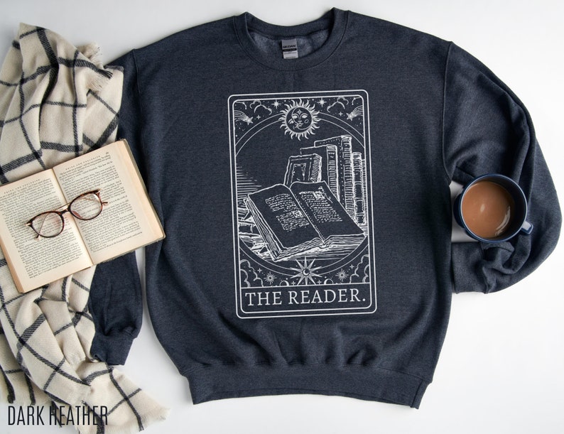 Puede incluir: Sudadera gris oscuro jaspeada con un gr&aacute;fico blanco de una carta de tarot que representa una pila de libros con el texto "THE READER."