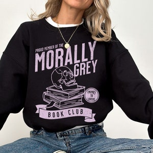 Puede incluir: Sudadera negra con un estampado gr&aacute;fico morado que dice "Proud Member of the Morally Grey Book Club". El gr&aacute;fico incluye una calavera con gafas, una pila de libros y un c&iacute;rculo que dice "Enemies to Lovers".