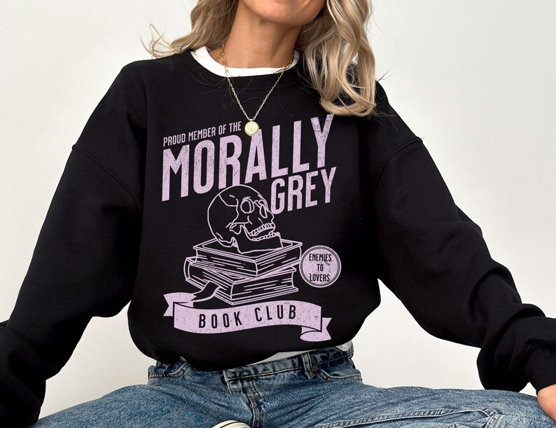 Puede incluir: Sudadera negra con un estampado gr&aacute;fico morado que dice "Proud Member of the Morally Grey Book Club". El gr&aacute;fico incluye una calavera con gafas, una pila de libros y un c&iacute;rculo que dice "Enemies to Lovers".