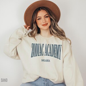 May include: A cream-colored sweatshirt with the text "Zodiac Academy Solaria" printed in blue on the front. The model is wearing a brown hat and blue jeans.