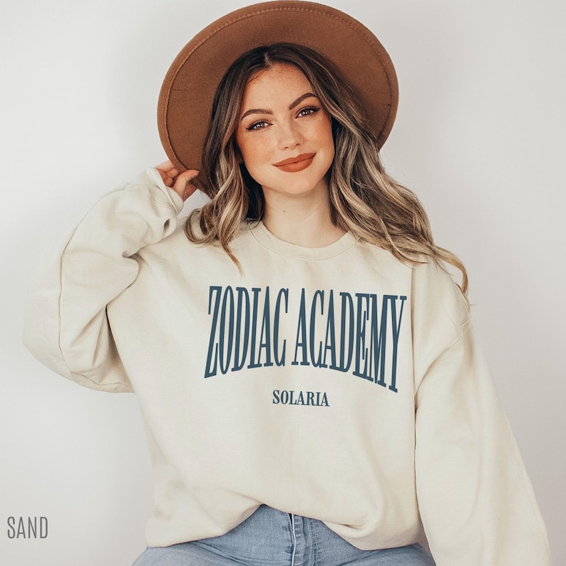 May include: A cream-colored sweatshirt with the text "Zodiac Academy Solaria" printed in blue on the front. The model is wearing a brown hat and blue jeans.