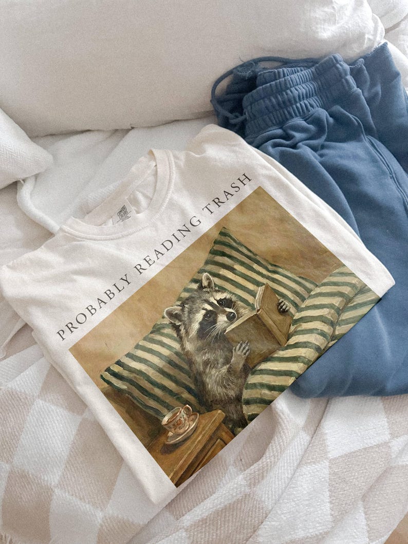 以下が含まれることがあります： 「PROBABLY READING TRASH」の文字と本を読むアライグマが描かれた白いTシャツ。青いスウェットパンツも写っており、チェック柄の毛布が敷かれたベッドの上に置かれています。