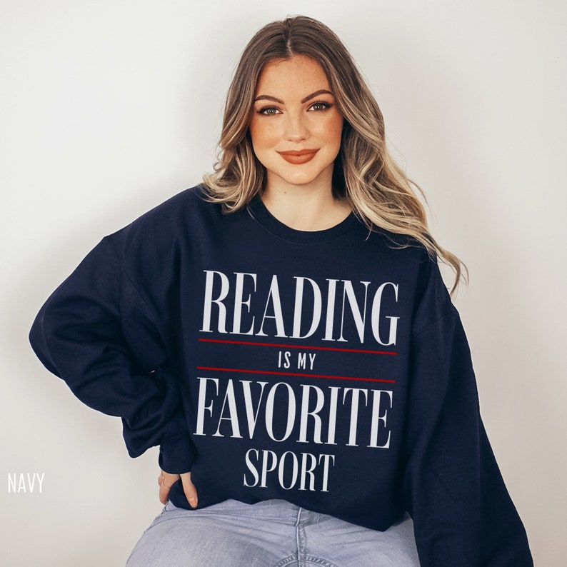 May include: Navy blue sweatshirt featuring the text "READING IS MY FAVORITE SPORT" in white, with a red line separating the phrases. A comfortable, casual style suitable for everyday wear.