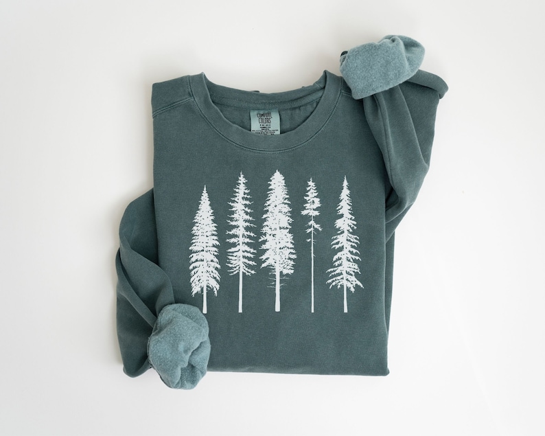 May include: A teal blue sweatshirt with a white graphic of five pine trees. The sweatshirt has a crew neck and long sleeves.