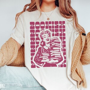 May include: A white t-shirt with a pink graphic of a woman holding a stack of books. The graphic has the text "BOOKISH" repeated in a grid pattern.