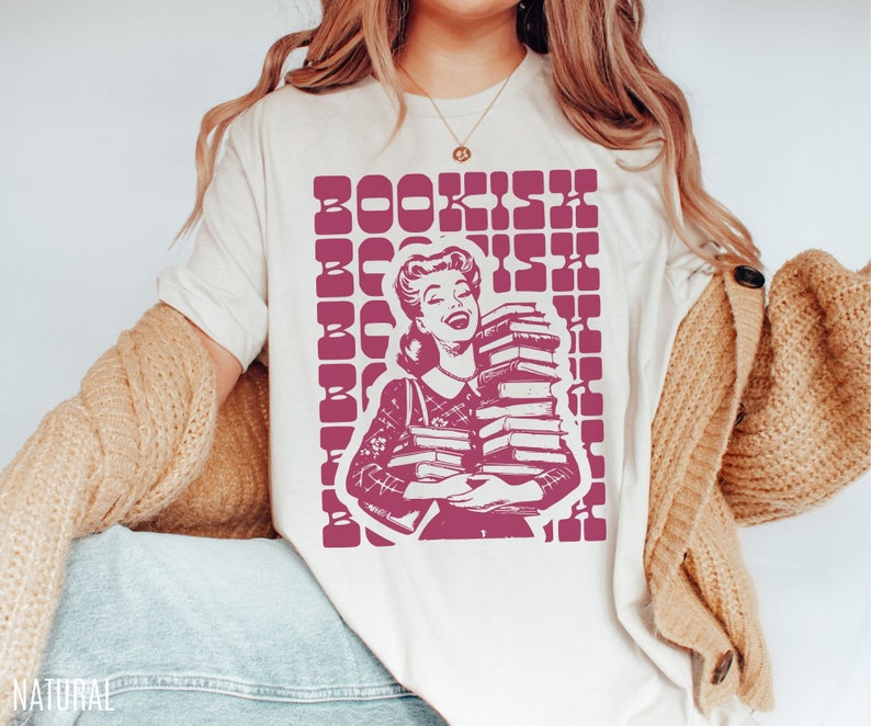 May include: A white t-shirt with a pink graphic of a woman holding a stack of books. The graphic has the text "BOOKISH" repeated in a grid pattern.