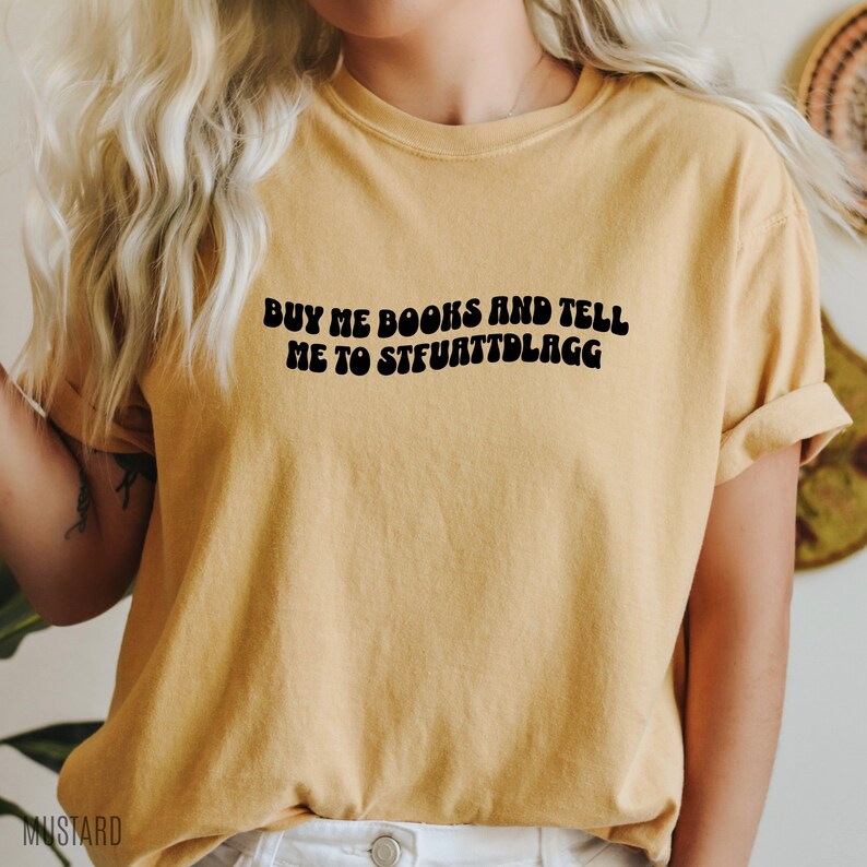 May include: Mustard-colored t-shirt with the text "BUY ME BOOKS AND TELL ME TO STFUATTDLAGG" in black. The shirt has short sleeves and a relaxed fit. The word "MUSTARD" is printed at the bottom left corner.