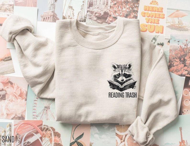 May include: Sand-colored sweatshirt featuring a black and white illustration of a raccoon reading a book. Below the raccoon, the text reads "READING TRASH". The sweatshirt has a crew neck and long sleeves.