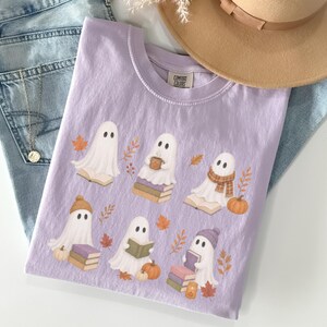 Orchid Comfort Colors shirt featuring a whimsical book lover ghost design with six reading ghosts, stacked books, pumpkins, fall foliage, knit hats, and a warm drink.
