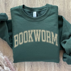 May include: Forest green sweatshirt with the word "BOOKWORM" in a distressed, arched font across the chest. The sweatshirt has a crew neck and ribbed cuffs. The garment is folded on a light wood surface.