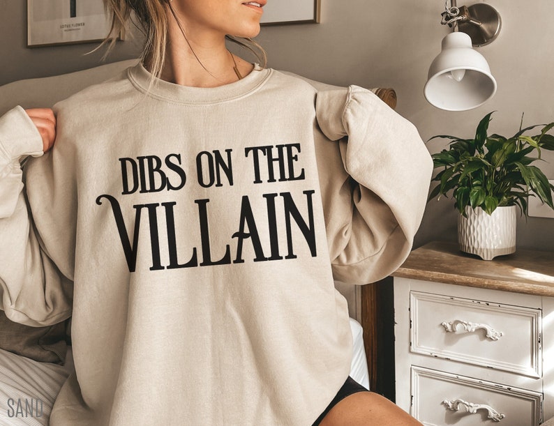 K&ouml;nnte beinhalten: Beiges Sweatshirt mit dem schwarzen Text "DIBS ON THE VILLAIN", eine verspielte und k&uuml;hne Aussage. Das Sweatshirt hat eine neutrale Farbe und eignet sich f&uuml;r Freizeitkleidung. Der Text ist in einer stilisierten Schriftart gehalten und verleiht dem Kleidungsst&uuml;ck eine pers&ouml;nliche Note.