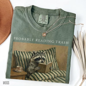 Folded moss green Comfort Colors t shirt with probably reading trash text and raccoon reading book design, styled with jewelry and dried leaves