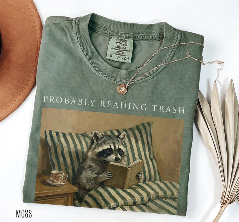 Folded moss green Comfort Colors t shirt with probably reading trash text and raccoon reading book design, styled with jewelry and dried leaves