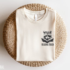 May include: A natural coloured t-shirt with a graphic of a raccoon reading a book and the text "Reading Trash".