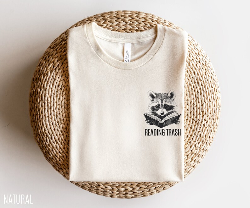 May include: A natural coloured t-shirt with a graphic of a raccoon reading a book and the text "Reading Trash".