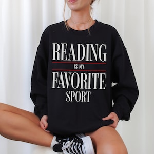 May include: Black sweatshirt featuring the text "READING IS MY FAVORITE SPORT" in white, with a red line separating the phrases. The sweatshirt is a crew neck style.