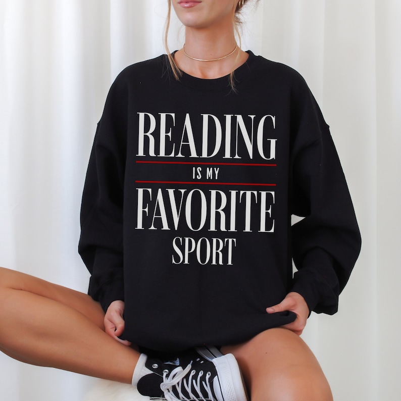 May include: Black sweatshirt featuring the text "READING IS MY FAVORITE SPORT" in white, with a red line separating the phrases. The sweatshirt is a crew neck style.