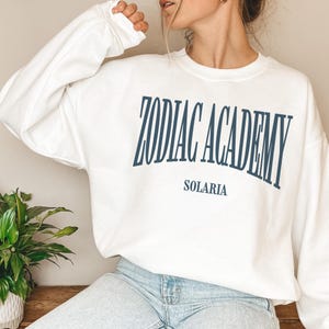 May include: White sweatshirt with the text "Zodiac Academy" in blue lettering and "Solaris" in smaller blue lettering below.