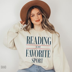 May include: A cream-colored sweatshirt with the text "Reading is my favorite sport" in navy blue and red. The model is wearing a brown hat and blue jeans.