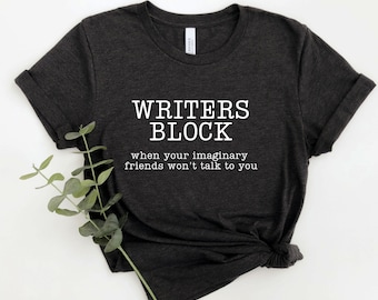 Könnte beinhalten: Dunkelgraues T-Shirt mit dem weißen Text "WRITERS BLOCK" und dem Satz "when your imaginary friends won't talk to you". Das T-Shirt ist gefaltet und hat einen Zweig mit grünen Blättern.