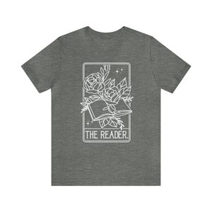 May include: A heather gray t-shirt featuring a white graphic of an open book with roses and leaves, framed by a rectangle. The text "THE READER" is below the image. The top of the frame has the Roman numeral "XII" and two small stars.