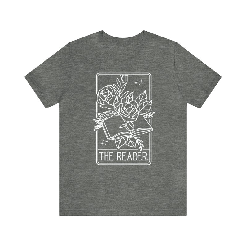May include: A heather gray t-shirt featuring a white graphic of an open book with roses and leaves, framed by a rectangle. The text "THE READER" is below the image. The top of the frame has the Roman numeral "XII" and two small stars.