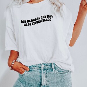 May include: White t-shirt with the text "BUY ME BOOKS AND TELL ME TO STFUATTLAGG" in black, curved font. The shirt is paired with light blue denim jeans. A bracelet is visible on the left wrist.