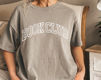 May include: A grey short-sleeved t-shirt featuring the text "BOOK CLUB" printed in a white collegiate-style font across the chest. The garment has a crew neckline and a relaxed fit, with a washed-out appearance to the fabric texture.