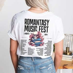 May include: White t-shirt featuring the text "ROMANTASY MUSIC FEST 2024" with a skull and floral design. The back of the shirt lists various romance novel tropes. A unique graphic tee for book lovers.