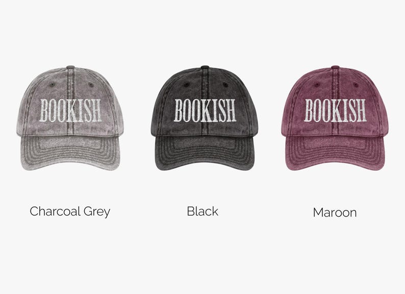 May include: Three baseball caps in charcoal grey, black, and maroon. Each cap has the word "BOOKISH" embroidered in white across the front. The caps are displayed against a white background.