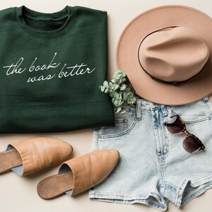 Puede incluir: Una sudadera de cuello redondo verde oscuro con el texto cursivo blanco "the book was better" impreso en el frente, junto con pantalones cortos de mezclilla azul claro, un sombrero beige de ala ancha, mules de cuero beige y gafas de sol de carey.