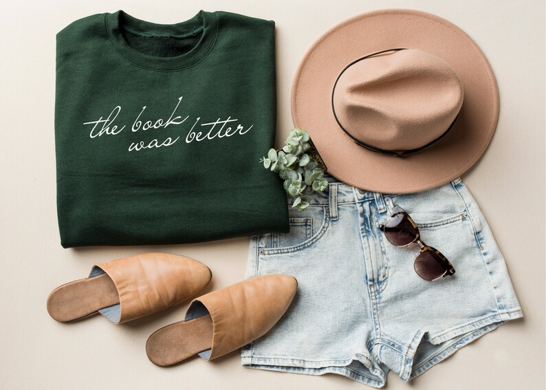 Puede incluir: Una sudadera de cuello redondo verde oscuro con el texto cursivo blanco "the book was better" impreso en el frente, junto con pantalones cortos de mezclilla azul claro, un sombrero beige de ala ancha, mules de cuero beige y gafas de sol de carey.