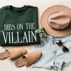 K&ouml;nnte beinhalten: Draufsicht auf ein l&auml;ssiges Outfit. Ein dunkelgr&uuml;nes Sweatshirt mit dem wei&szlig;en Text "DIBS ON THE VILLAIN" wird mit hellblauen Jeansshorts, einem beigefarbenen Fedora-Hut, einer Sonnenbrille und beigefarbenen Lederslippern kombiniert. Ein Zweig Gr&uuml;nzeug ist ebenfalls vorhanden.