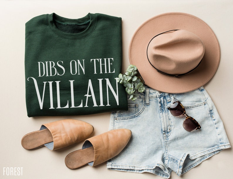 K&ouml;nnte beinhalten: Draufsicht auf ein l&auml;ssiges Outfit. Ein dunkelgr&uuml;nes Sweatshirt mit dem wei&szlig;en Text "DIBS ON THE VILLAIN" wird mit hellblauen Jeansshorts, einem beigefarbenen Fedora-Hut, einer Sonnenbrille und beigefarbenen Lederslippern kombiniert. Ein Zweig Gr&uuml;nzeug ist ebenfalls vorhanden.