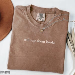 May include: Espresso-colored t-shirt with the phrase "will yap about books" in white. The shirt has a crew neck and short sleeves. A silver necklace with a small pendant rests on the shirt. The shirt is folded on a white surface.