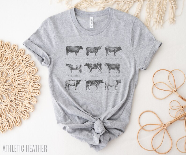 Puede incluir: Camiseta gris jaspeado con una ilustraci&oacute;n detallada de varias razas de vacas, incluyendo Black Angus, Highland y Brown Swiss. El dise&ntilde;o de la camiseta incluye los nombres de cada raza. El texto "ATHLETIC HEATHER" est&aacute; en la parte inferior.
