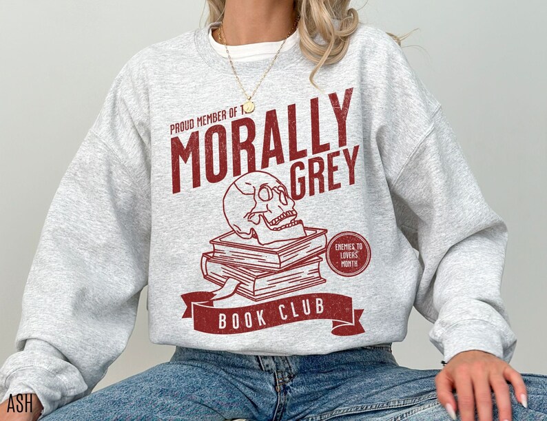 Puede incluir: Sudadera gris con un dise&ntilde;o gr&aacute;fico rojo que dice "Proud Member of the Morally Grey Book Club". El gr&aacute;fico tambi&eacute;n incluye una calavera y huesos cruzados y el texto "Enemies to Lovers Month".