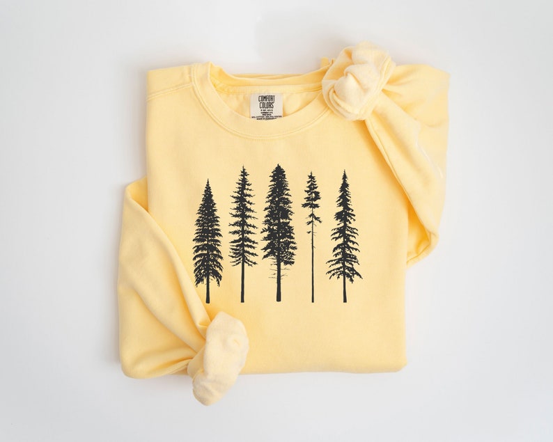 May include: A yellow sweatshirt with a black silhouette of five pine trees printed on the front. The sweatshirt is folded in half and the sleeve is rolled up.