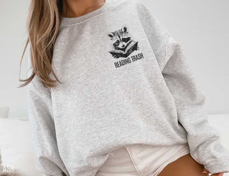 May include: Light gray sweatshirt featuring a raccoon reading a book, with the text "READING TRASH" printed below. The raccoon illustration is in black and white. The sweatshirt is a casual style.