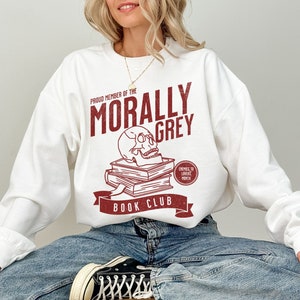 Puede incluir: Sudadera blanca con un dise&ntilde;o gr&aacute;fico rojo que dice "Proud Member of the Morally Grey Book Club". El gr&aacute;fico incluye una calavera con libros apilados encima y un c&iacute;rculo rojo que dice "Enemies to Lovers Month".