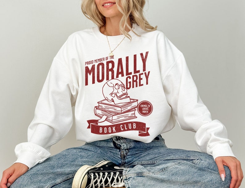 Puede incluir: Sudadera blanca con un dise&ntilde;o gr&aacute;fico rojo que dice "Proud Member of the Morally Grey Book Club". El gr&aacute;fico incluye una calavera con libros apilados encima y un c&iacute;rculo rojo que dice "Enemies to Lovers Month".