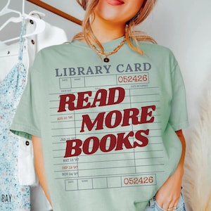 Woman wearing a bay green Comfort Colors t shirt featuring a nostalgic library card illustration with bold red Read More Books typography, styled with layered gold necklaces and denim for a relaxed reading inspired outfit
