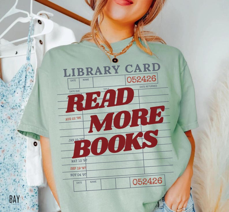 Woman wearing a bay green Comfort Colors t shirt featuring a nostalgic library card illustration with bold red Read More Books typography, styled with layered gold necklaces and denim for a relaxed reading inspired outfit