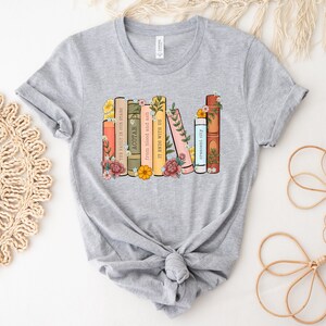 May include: Gray t-shirt with a graphic of a stack of books with floral accents. The book titles include "The Fault in Our Stars", "ACOTAR", "from blood and ash", "IT ENDS WITH US", and "crescent city".