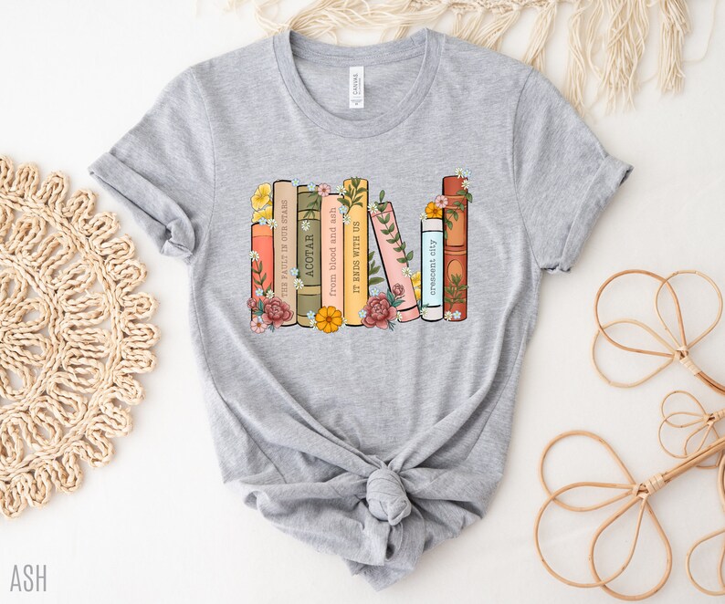 May include: Gray t-shirt with a graphic of a stack of books with floral accents. The book titles include "The Fault in Our Stars", "ACOTAR", "from blood and ash", "IT ENDS WITH US", and "crescent city".