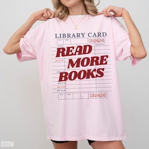 Woman holding an oversized blossom pink Comfort Colors t shirt featuring a retro library card graphic and bold Read More Books text, styled with black biker shorts and gold jewelry for a cozy bookworm aesthetic
