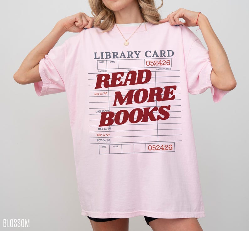 Woman holding an oversized blossom pink Comfort Colors t shirt featuring a retro library card graphic and bold Read More Books text, styled with black biker shorts and gold jewelry for a cozy bookworm aesthetic