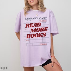 Woman wearing an oversized orchid Comfort Colors t shirt with a retro library card design and bold red Read More Books text, styled with black biker shorts and gold jewelry for a cozy book lover aesthetic